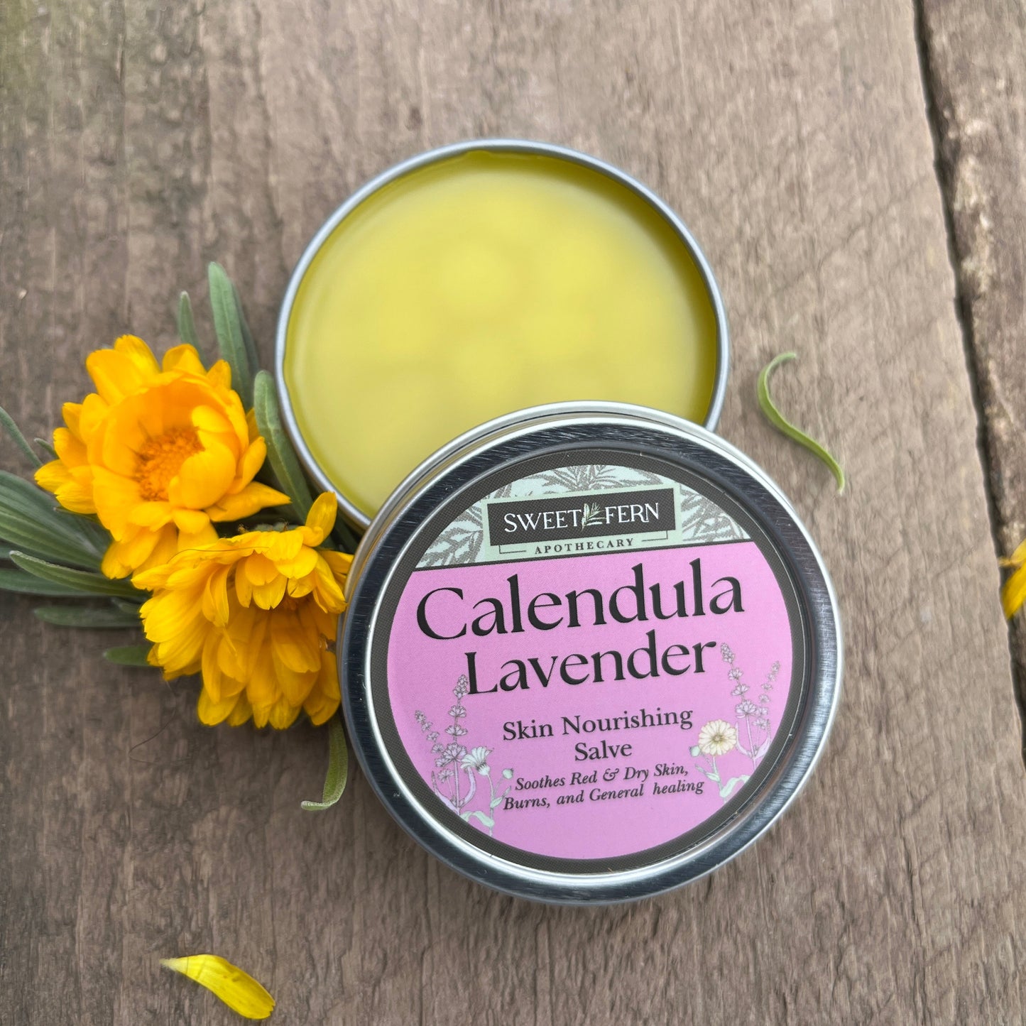 Open tin of Sweet Fern Apothecary Calendula Lavender Skin Nourishing Salve on a wooden surface with yellow flowers.